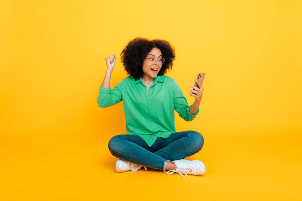 Foto a figura intera di eccitata felice donna afro-americana o brasiliana riccia, si siede su uno sfondo giallo, utilizza il suo smartphone, navigando in internet, gioisce per le notizie, gesticolando mani, sorrisi - Foto, immagini