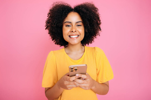 Ładna, hiszpańska lub brazylijska kobieta, w żółtej sukience, korzystająca ze smartfona, rozmawiająca online z przyjaciółmi, przeglądająca internet, smsująca, patrząca w kamerę, uśmiechnięta, stojąca na odizolowanym różowym tle - Zdjęcie, obraz