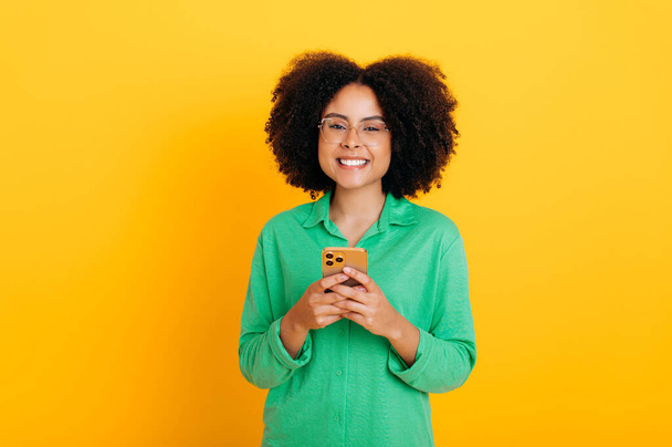 Positive beautiful brazilian or african american curly haired young woman wearing green shirt, with glasses, holding her smartphone, looking at camera, smiling, stand on isolated yellow background - Fotografie, Obrázek
