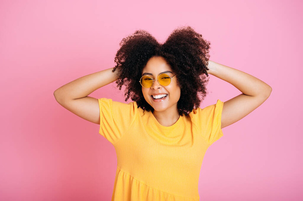 Encantadora mujer afro-americana emocionada alegre o hispana de pelo rizado en gafas y vestido naranja de moda, divirtiéndose, posando, tomándose de la mano en el cabello, mira a la cámara, sonrisa, fondo rosa aislado - Foto, imagen
