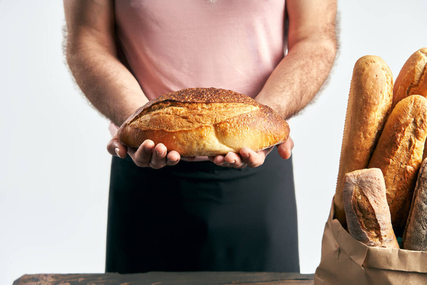 Baker man in apron holding loaf of fresh baked bread in the hands. Bakery, food concept - Фото, зображення