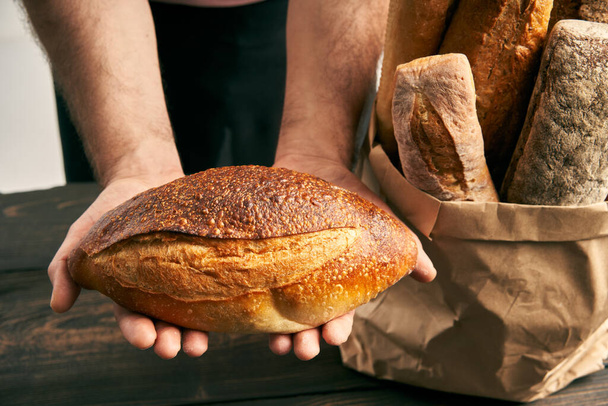 Baker man in apron holding loaf of fresh baked bread in the hands. Bakery, food concept - Foto, Imagem
