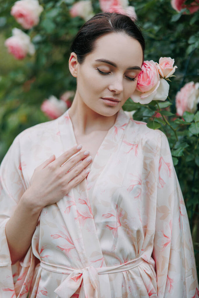 Portrait of young brunette woman in kimono dress with closed eyes enjoying by blooming pink Eden rose in garden. - Foto, Bild