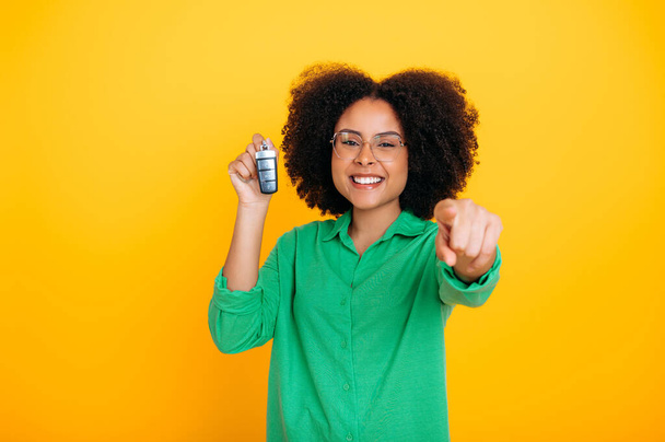 Car for you. Happy african american or brazilian beautiful woman with curly hair, in green shirt, holding car key in hand, rejoices in new purchase, smile at camera,stand on isolated orange background - Φωτογραφία, εικόνα