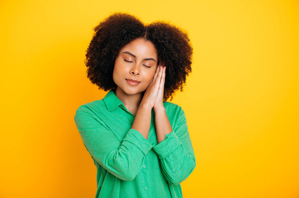 Beautiful calm brazilian or african american curly haired woman, in a green shirt, makes a sleep gesture with her palms, closed her eyes, ready to fall asleep, isolated yellow background - Foto, Imagem