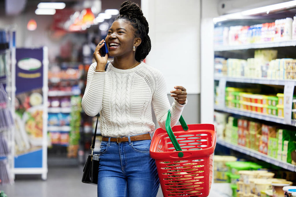 Happy black woman, supermarket and shopping with phone call, choice or discount in funny chat, customer experience. Plastic basket, grocery store and mall for retail, talking and girl with smartphone. - Zdjęcie, obraz