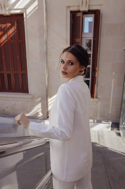 fashion outdoor photo of beautiful sensual woman with dark hair in luxurious fashion outfit, elegant white pants and jacket, with accessories - Valokuva, kuva
