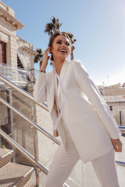 fashion outdoor photo of beautiful sensual woman with dark hair in luxurious fashion outfit, elegant white pants and jacket, with accessories - 写真・画像
