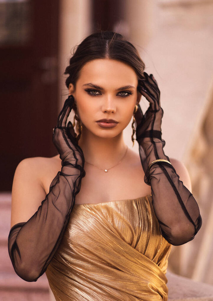 fashion outdoor photo of beautiful sensual woman with dark hair in luxurious fashion outfit, elegant golden dress with black gloves and accessories, posing in front of building in classic style - Fotoğraf, Görsel