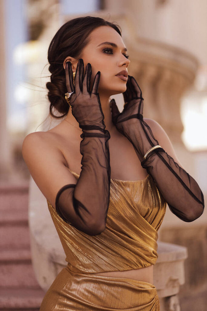 fashion outdoor photo of beautiful sensual woman with dark hair in luxurious fashion outfit, elegant golden dress with black gloves and accessories, posing in front of building in classic style - Fotoğraf, Görsel