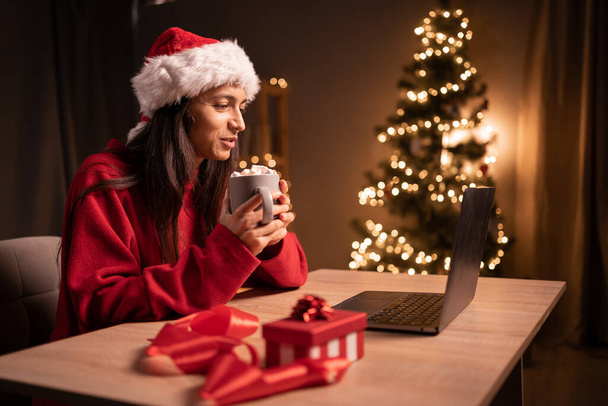 Νεαρή ευτυχισμένη γυναίκα που έχει βιντεοκλήση πάνω από το φορητό υπολογιστή, ενώ γιορτάζει τα Χριστούγεννα μόνη στο σπίτι. Δες ταινία με ζαχαρωτά. Αντιγραφή χώρου - Φωτογραφία, εικόνα