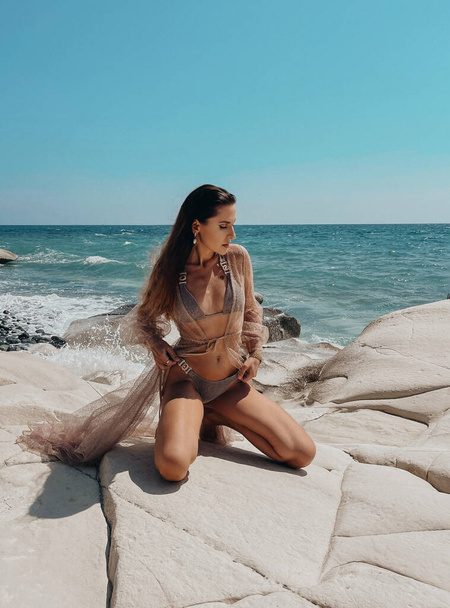 fashion outdoor photo of beautiful woman with dark hair in elegant swimming suit posing at the rocky beach - Zdjęcie, obraz