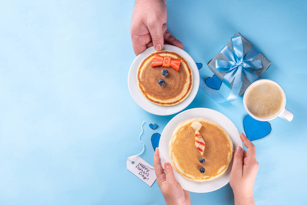 Father's day holiday idea, cute surprise gift. Child preparing breakfast for dad, creative decorated pancakes with tie, bow tie, inscription I love Dad. On light blue background, with child, dad hands - Photo, Image