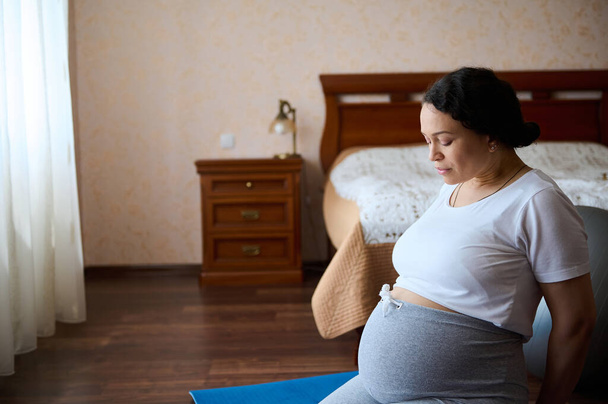 Egy középkorú terhes nő portréja, akinek nagy hasa van a terhesség harmadik harmadában, és otthon edz egy jóga szőnyegen. Gyereket várok. Terhesség. Anyaság és anyaság életmód fogalma - Fotó, kép