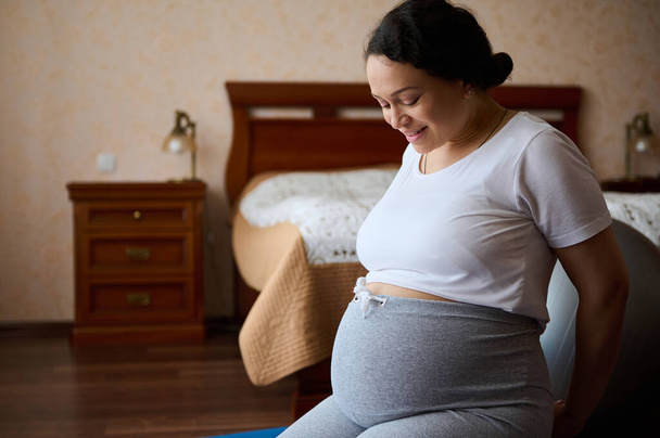 Happy adult pregnant woman, expectant mom sitting on floor, smiling looking at her big belly in late pregnancy, feeling baby movements, smiling enjoying her pregnancy and contact with her future child - Photo, Image