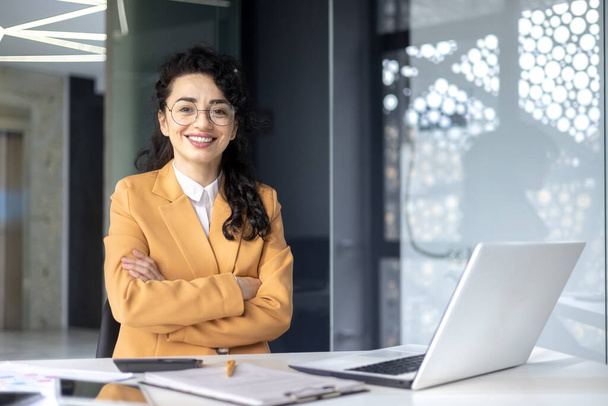Portrait of successful business woman boss in yellow suit, mature financier marketer at desk smiling and looking at camera with crossed arms, female worker using laptop at work. - Zdjęcie, obraz