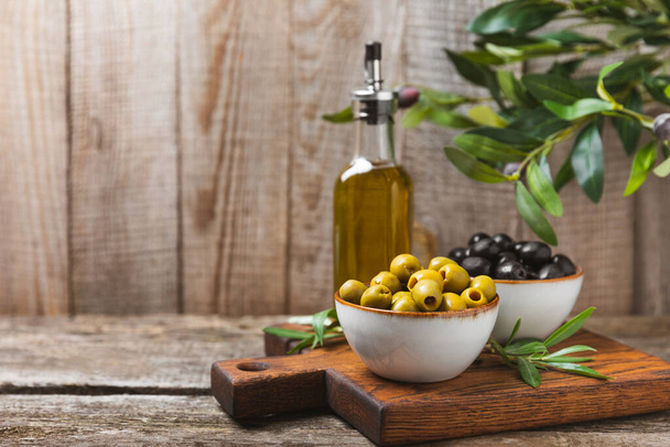 Green and black olives on a brown wooden background. Various types of olives in bowls and olive oil with fresh olive leaves. Copy space. Place for text. Mediterranean food. Vegan. - Photo, image