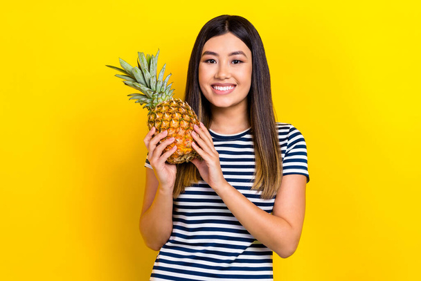 Portrait of young funky vietnamese woman hold ananas eat exotica fruit wear striped t-shirt vitamins isolated on yellow color background. - 写真・画像