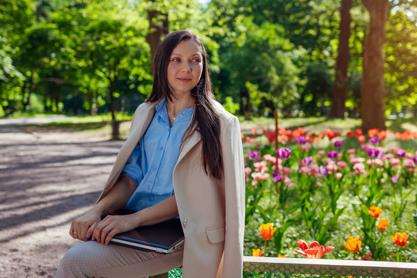 Venkovní portrét mladé podnikatelky pracující v jarním parku na notebooku. Žena krajinář má výsledky kvetoucích květinových záhonů ve veřejné zahradě - Fotografie, Obrázek