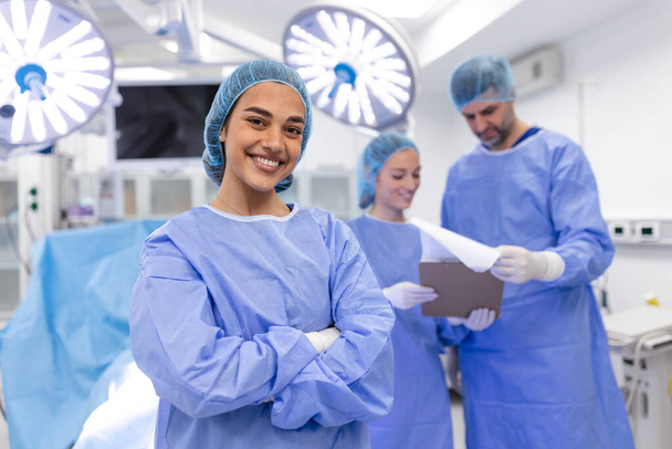 Detailní záběr chirurgky, jak se dívá na kameru s kolegy vystupujícími v zázemí operačního sálu. Koncept medicíny - Fotografie, Obrázek