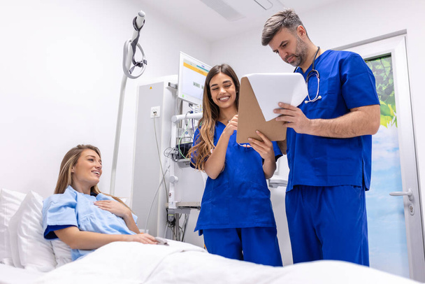Lékaři v uniformě se stetoskopem používají podložku, která stojí na nemocničním oddělení a pacient odpočívá v posteli na pozadí. Lékař kontroluje údaje o pacientech - Fotografie, Obrázek
