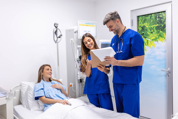 Lékaři v uniformě se stetoskopem používají podložku, která stojí na nemocničním oddělení a pacient odpočívá v posteli na pozadí. Lékař kontroluje údaje o pacientech - Fotografie, Obrázek