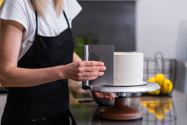 Pastelero profesional que utiliza raspador de pastelería para manejar la almáciga blanca en la mujer de la torta horneada en delantal negro disfrutando del proceso de preparación de pasteles personalizados orgánicos en casa - Foto, imagen