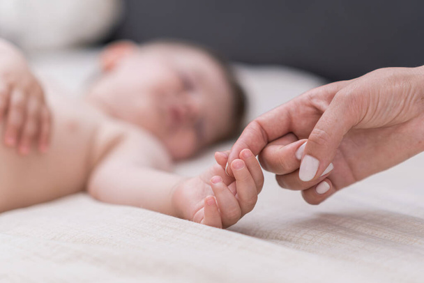 Śpiący mały chłopiec chwytający palec matki z malutką rączką leżącą na miękkim łóżku z białą prześcieradłem kobieta wyrażająca miłość i uczucie do syna w sypialni zbliżenie - Zdjęcie, obraz