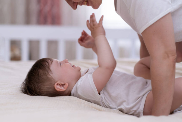 Cheerful baby boy stretching out hands to touch face of mommy lying on soft bed in nursery happy parent showing love and entertaining little son in morning at home - Foto, Imagem
