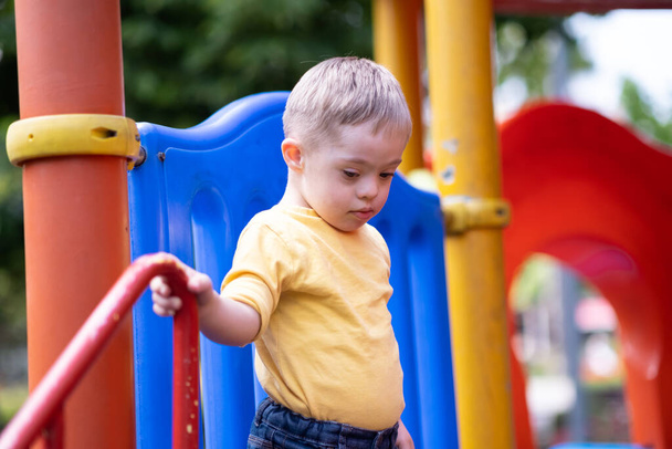 Netter blonder Junge mit Down-Syndrom steht oben und bereitet sich auf das Reiten der bunten Rutsche vor. Kleines Kind genießt Unterhaltung auf dem modernen Spielplatz einer Wohnanlage in der Stadt - Foto, Bild