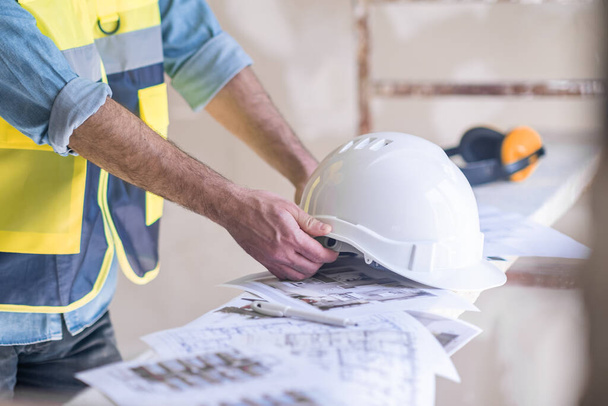Skilled worker in uniform taking white protective helmet lying on heap of project papers specialist getting ready for renovation in room construction safety rules and equipment - Photo, Image