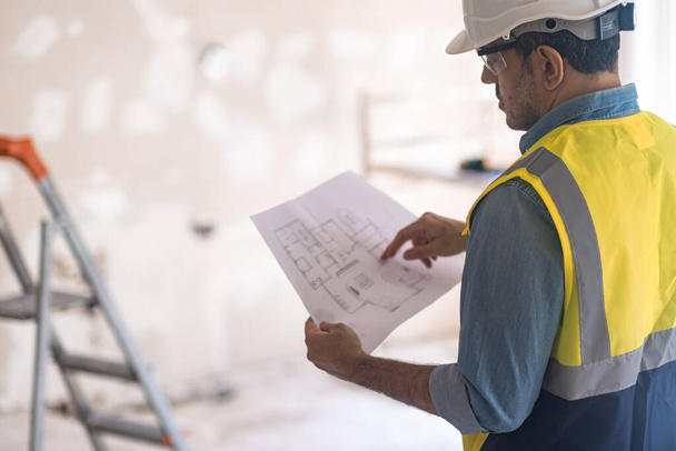 Inżynier w profesjonalnym mundurze trzymający plan projektu mieszkania sprawdza szczegóły przed wykonaniem remontu pracownik w kamizelce trzymający papier w rękach stoi na drabinie w pomieszczeniu - Zdjęcie, obraz