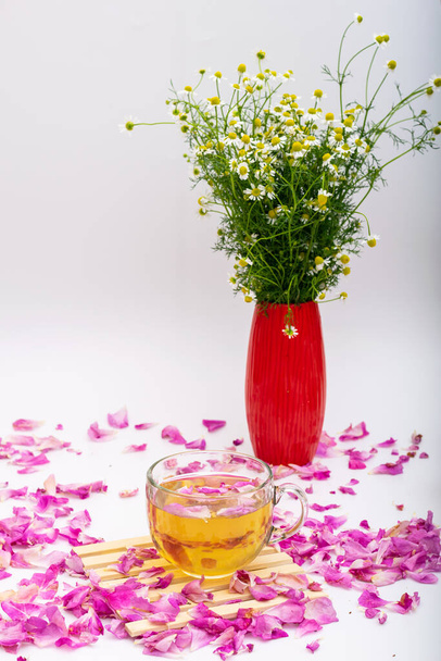 Herbal tea in glass cup on edible rose petals, vase with chamomile flowers, still life - Photo, Image