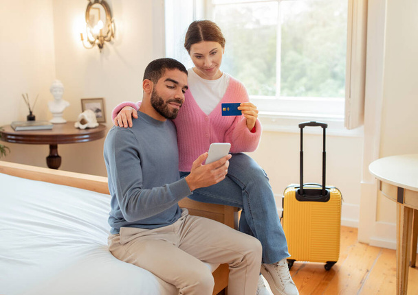 Bank Offer. Tourists Couple Booking Hotel Room Using Credit Card And Mobile Phone Sitting In Bedroom Indoors. Travelers Shopping Online In Application. Easy And Safe Payment Offer - Fotografie, Obrázek