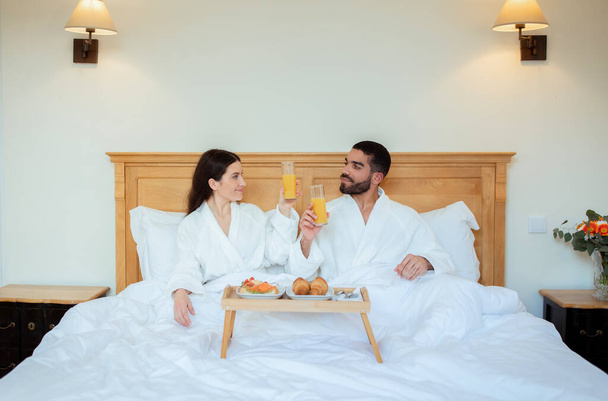 Casal alegre que aprecia o café da manhã, vidros de Clinking com suco de laranja que relaxa na cama, vestindo roupões de banho brancos no quarto confortável do hotel dentro de casa. Conceito de Vibrações de Férias de Lua de Mel - Foto, Imagem