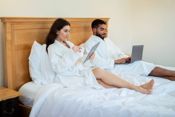 Aletler. Otelin iç kısmında, dizüstü bilgisayar ve tablet bilgisayar kullanan, beyaz bornoz giyen bir çiftin yan görüntüsü. Eş Yatak Odasında İnternette Sörf Yapıyor, Birlikte Çalışıyor - Fotoğraf, Görsel