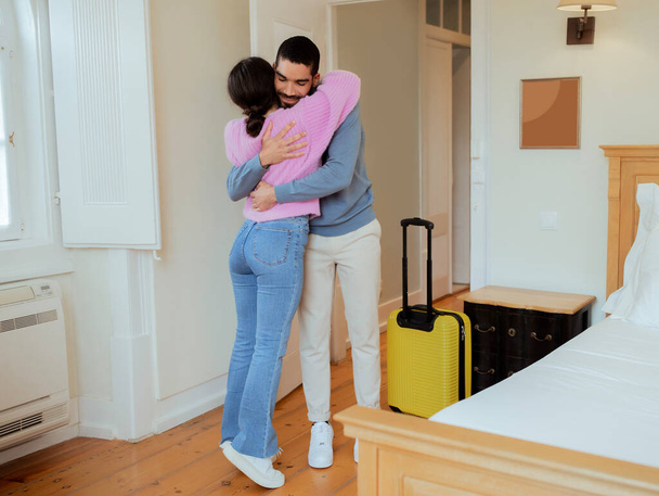 Honemoon Romance. Loving Spouses Hugging Standing With Suitcase At Hotel Suite Interior. Young Middle Eastern Couple Celebrating Romantic Vacation. Luxurious Accomodation Concept - 写真・画像