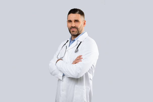 Medical services concept. Portrait of middle aged man doctor therapist in uniform posing with folded arms and looking at camera over light studio background, copy space - Foto, imagen