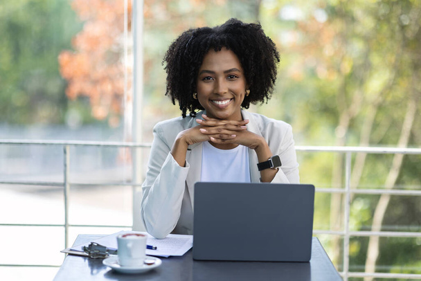 Pretty young black lady wearing smart casual outfit businesswoman sitting at cafe, using laptop, drinking coffee, working on business project, looking at camera and smiling, copy space - Foto, Imagem