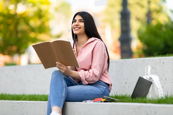 Pretty happy hispanic lady holding pen writing in notebook while sitting in park with laptop computer, dreamy female thinking, planning future - Foto, Bild