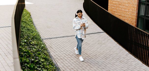 Ευτυχισμένη έφηβη κυρία κουβεντιάζοντας στα κοινωνικά δίκτυα στο κινητό και φορώντας ακουστικά ενώ περπατάτε στην αστική περιοχή της πόλης, πανόραμα, αντιγραφή χώρου - Φωτογραφία, εικόνα