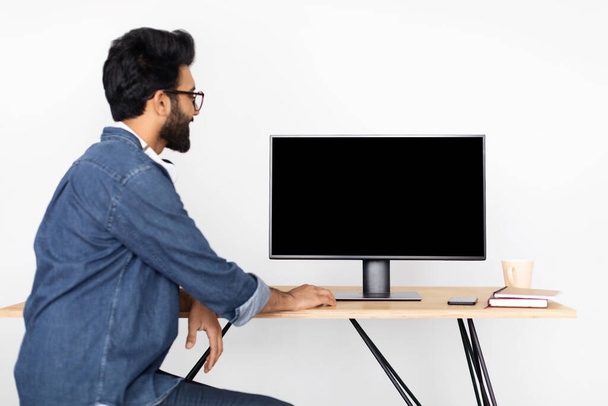 Ближневосточный бородатый молодой человек в очках сидит за рабочим столом, используя компьютер с чистым черным экраном, программист работает в офисе, белый фон стены, вид сзади, макет - Фото, изображение
