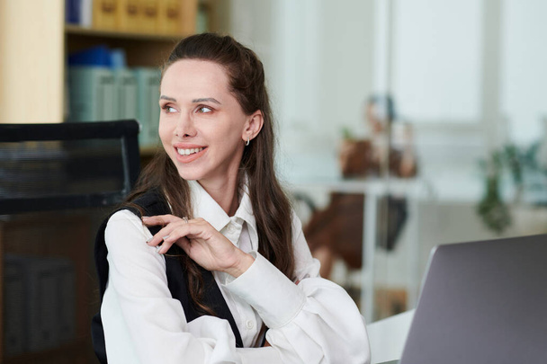 Νεαρή χαμογελαστή επιχειρηματίας που κάθεται στο χώρο εργασίας της με φορητό υπολογιστή στο γραφείο - Φωτογραφία, εικόνα