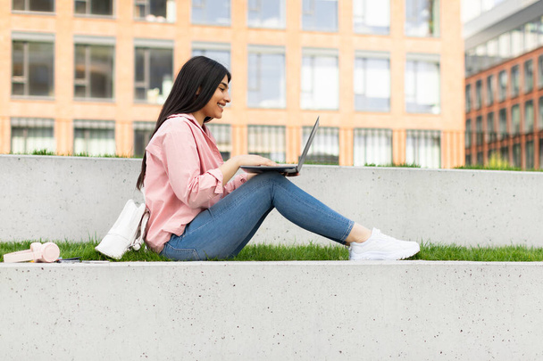 Девушка-подросток, использующая ноутбук на открытом воздухе, веселая женщина, обучающаяся онлайн, сидящая в студенческом городке и готовящаяся к экзаменам, боковой вид, свободное пространство. Счастливый студент, обучающийся дистанционно - Фото, изображение