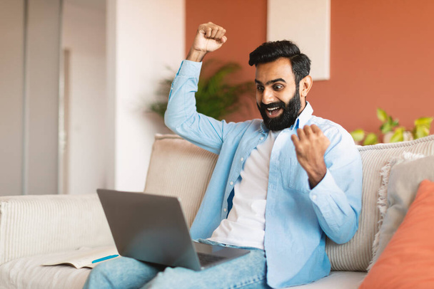 Ja. Freudiger Mann aus dem Nahen Osten mit Laptop und erhobenen Fäusten vor Freude und Feier im modernen Wohnzimmer sitzend. Indian Guy feiert große Online-Nachrichten und Glück, schreit Ja zu Hause - Foto, Bild
