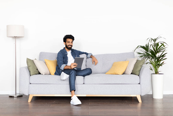 Positive happy handsome bearded arab guy wearing casual outfit sitting on couch at home over white background, have online meeting with lover, using digital tablet, copy space. Telecommunication - Фото, изображение