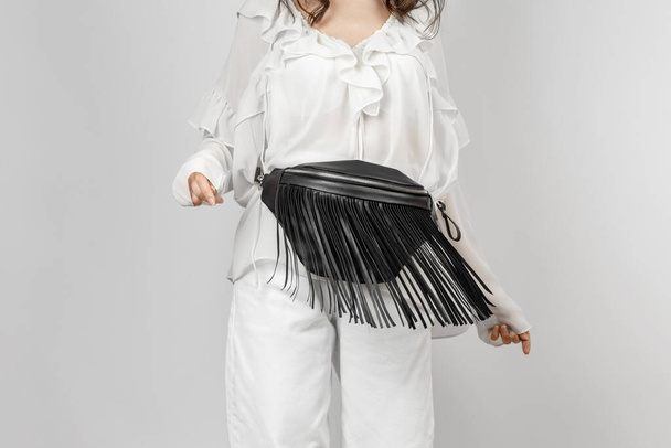 Valkoinen nainen yllään valkoiset farkut, pusero Frills ja Crossbody musta nahkavyö laukku koriste Fringe yli harmaa tausta - Valokuva, kuva