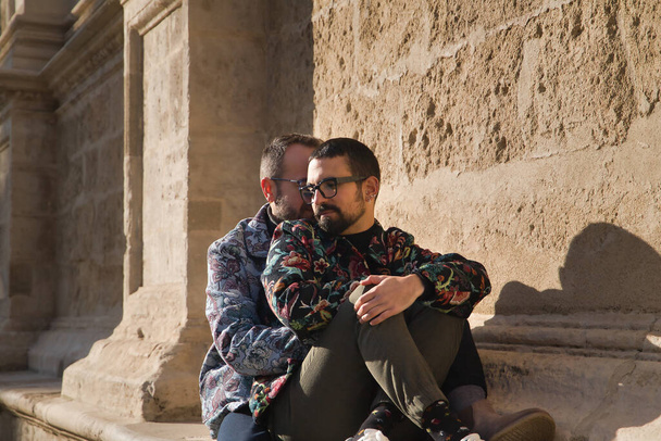 Real marriage of gay couple, sitting on a stone wall while one embraces the other from behind with very affectionate, complicit and happy attitude. Concept lgtb, lgtbiq+, couples, in love, love. - Valokuva, kuva