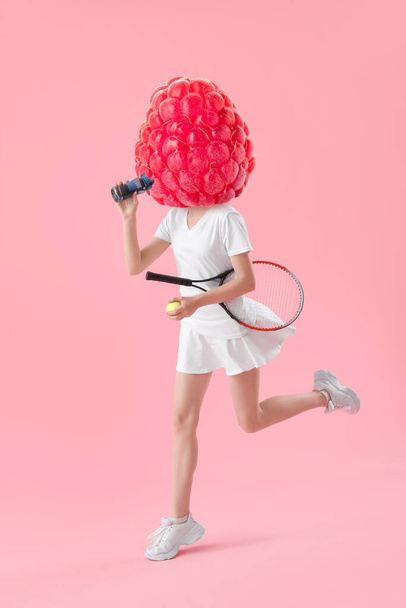 Running female tennis player with bottle of water and fresh raspberry instead of her head on pink background - Photo, Image