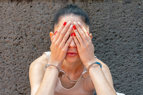 Eine Frau in Handschellen bedeckt ihr Gesicht mit den Händen gegen eine graue Wand. Konzept: Verhaftung und Inhaftierung, schöner Krimineller. - Foto, Bild